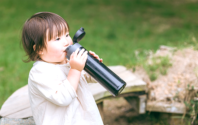 緑の芝生を背景に自分で大きな水筒を飲んでいる女児の画像