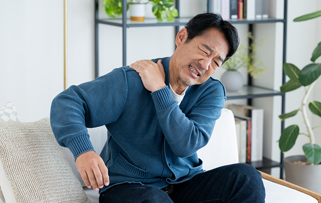 肩が凝って痛みを訴える働き盛りのソファに座る男性の画像