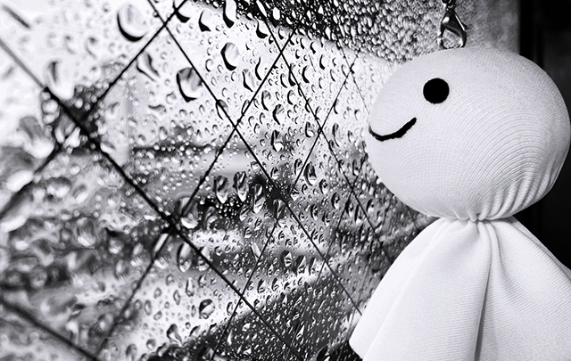 テルテル坊主がが窓ガラスに流れる雨のしずくを眺めている様子の画像