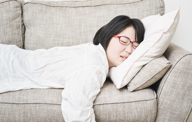 体調不良でソファに積まれたクッションにうつぶれになる女性の画像