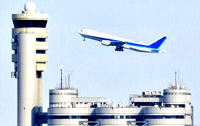 飛行場の管制塔とその向こうを飛ぶ飛行機の画像