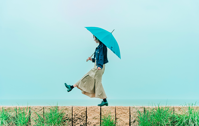 明るい雨の日に石段の上を水色の傘をさして足を上げて楽しそうに歩く女性の画像