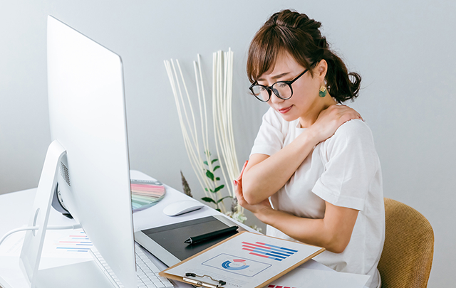 肩を抑えながら疲れをとろうとしているパソコンの大きなモニター前で仕事をする若い女性の画像