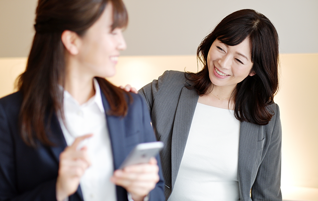 スマホに夢中にあている同僚に肩に手をかけ挨拶する２人の女性の画像