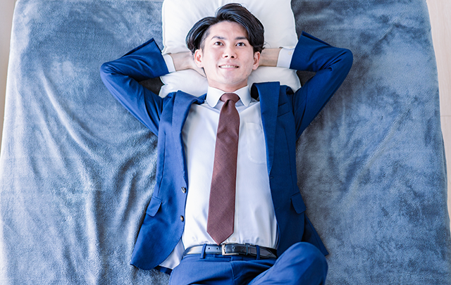 いつでも起きれる体制で、スーツを着たままベッドに横になるビジネスパーソンを天井から見下ろした画像