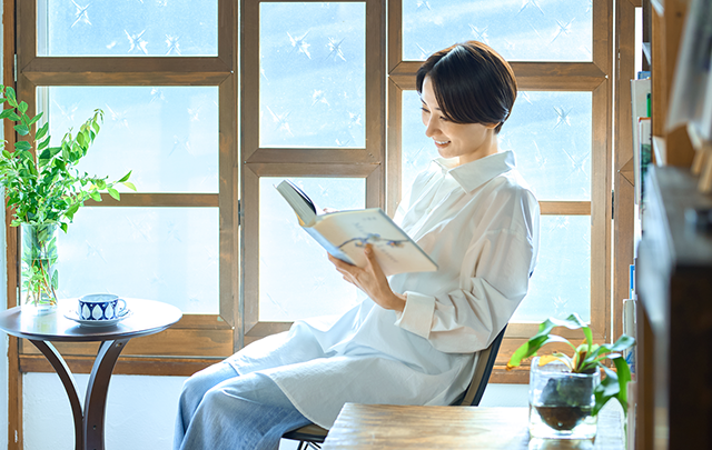 カフェのテラス側の窓辺で読書し、ゆとり時間を楽しむ女性の画像