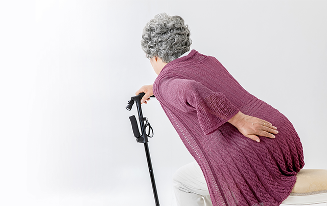 フレイルで腰が曲がった女性高齢者が杖をついて背中を抑えている画像