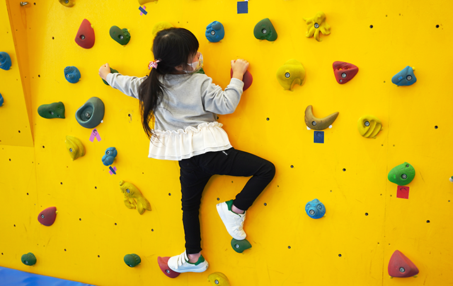 公園の真黄色の壁面のボルダリング遊具を登る小学生低学年の女児の画像
