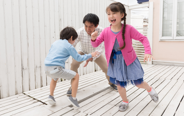 お父さんと小学生の姉弟の３人でテラスで鬼ごっこをしているほほえましい家族の画像