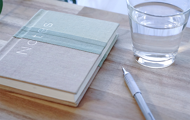 窓の前のデスクに閉じられたノートブック（日記帳）とペンと飲みかけのコップが置かれた画像