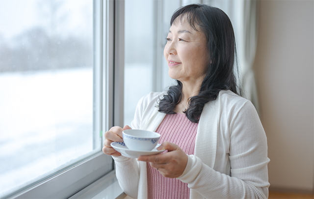 中年女性がコーヒカップを持って窓から雪景色を見ている画像