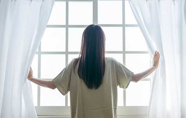 窓に向かって朝のカーテンを開けて陽を浴びる女性の画像